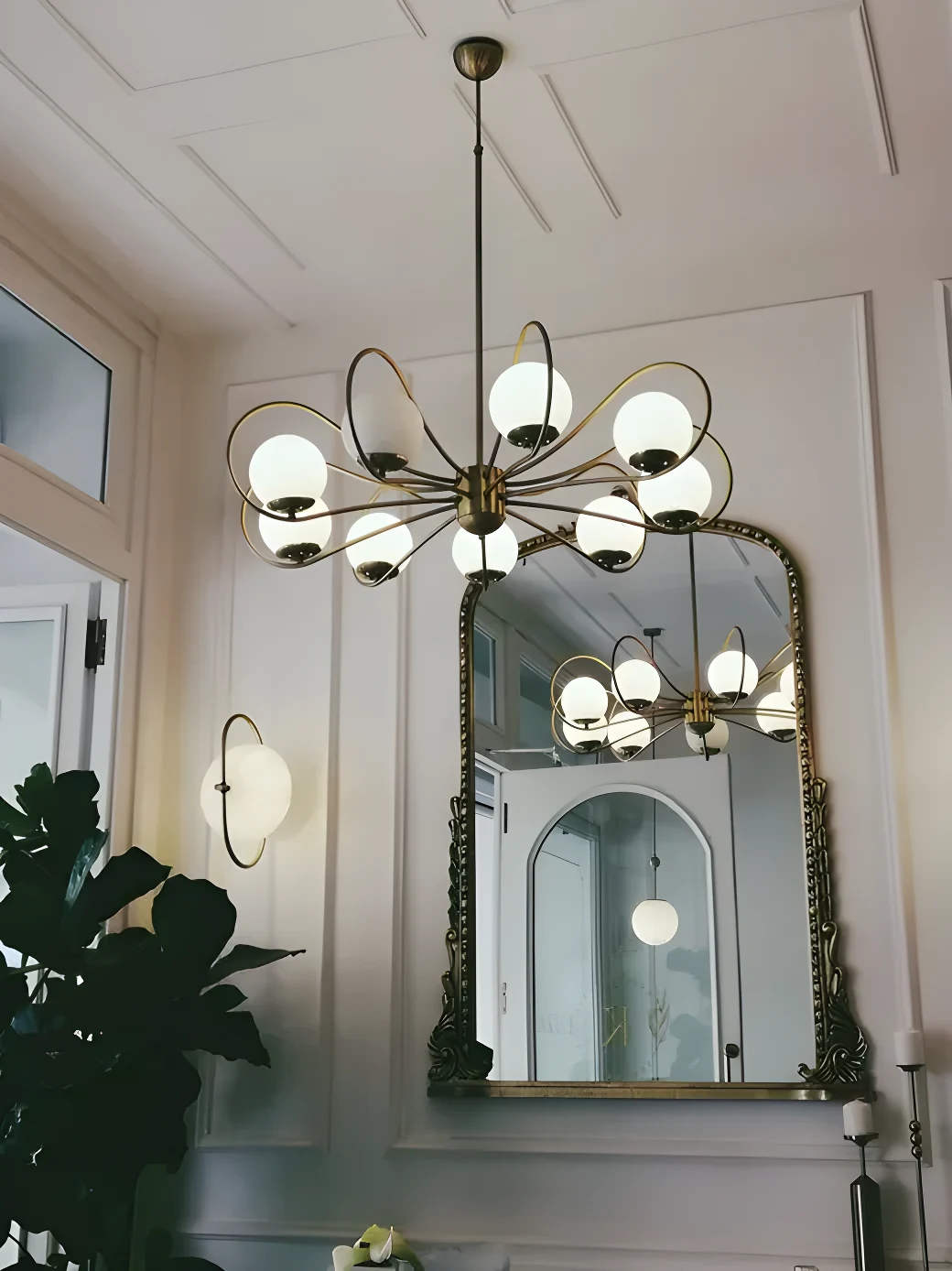 Lampadario in ottone con bracci curvi e sfere in vetro opalino, applique abbinata e grande specchio con cornice dorata intagliata su una parete bianca con boiserie