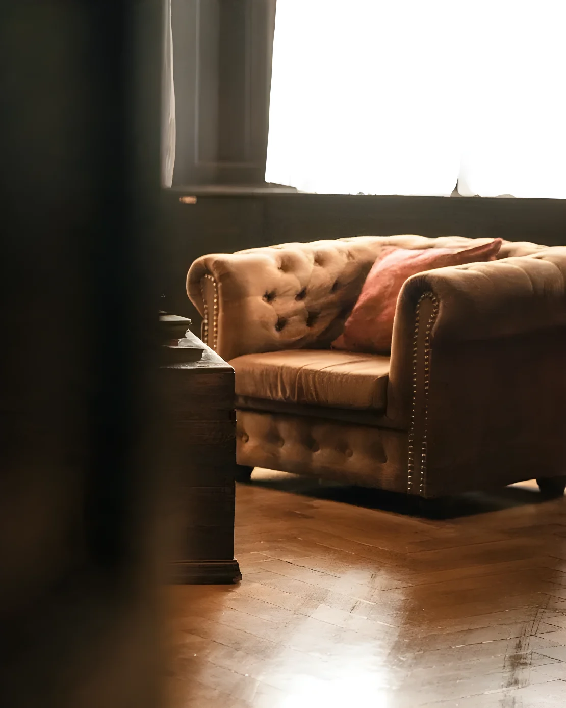 Poltrona Chesterfield in velluto color cammello con borchie e cuscino rosso, accanto a una cassapanca in legno scuro su parquet a spina di pesce, in controluce davanti a una finestra